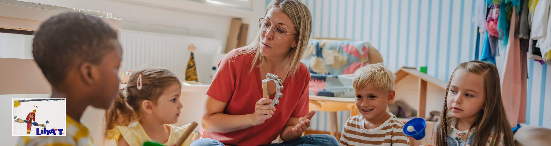 A teacher or caregiver in a red shirt sits with four young children in a classroom or daycare setting, engaging with them while holding an object. Colorful toys and striped wallpaper are visible in the background.
