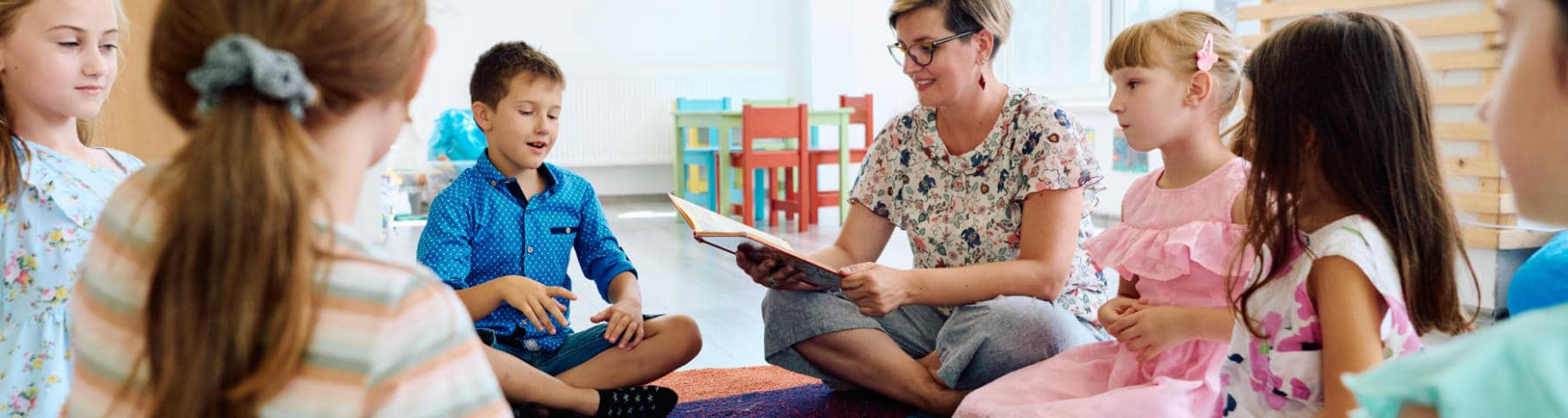 A female teacher with glasses sits on the floor in a circle with a diverse group of young children, reading from a book in a brightly lit classroom.
