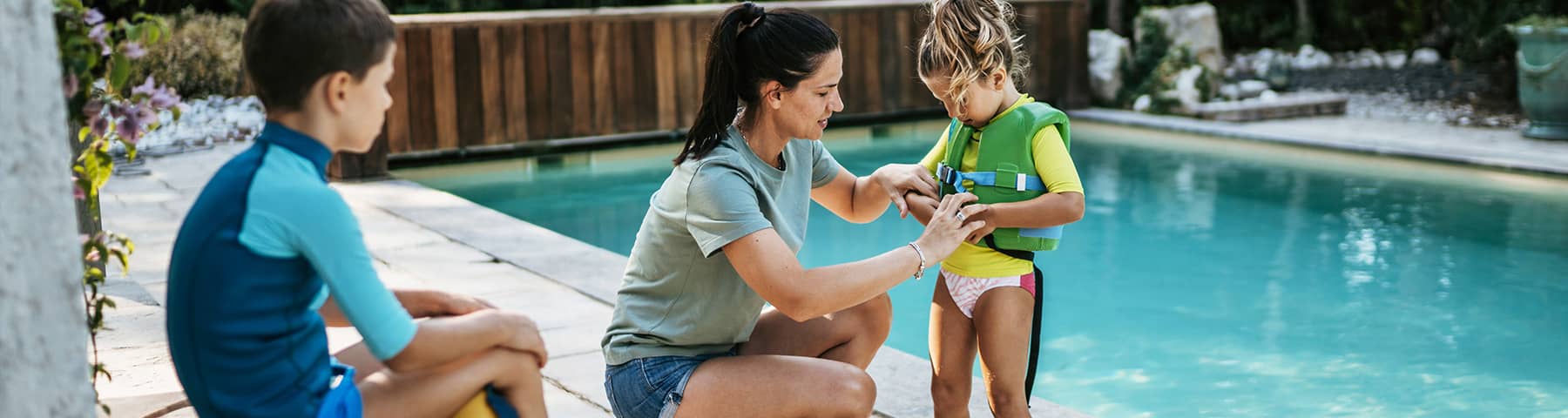 A woman kneels by a swimming pool helping a young child put on a green life jacket, while another child in a blue swimsuit sits nearby watching.