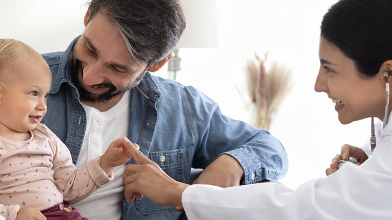 A smiling doctor with a stethoscope talks to a father holding a baby during a pediatric checkup.