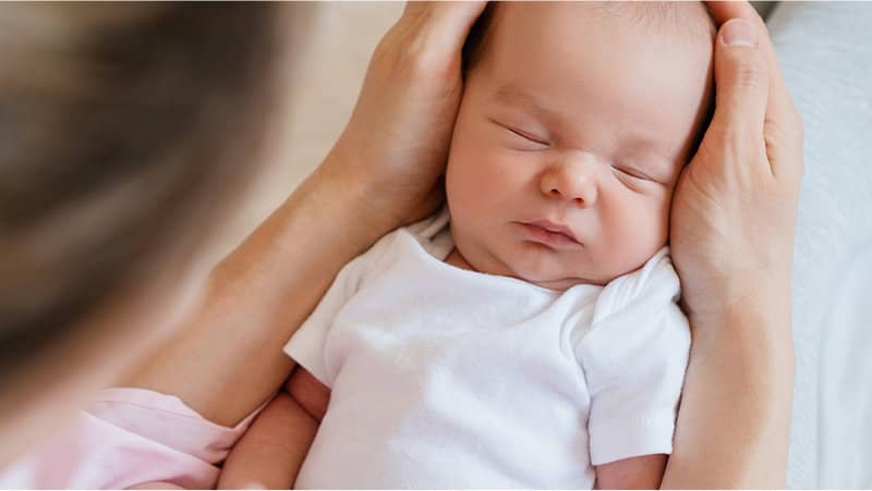 Close-up of a sleeping newborn in a white onesie being gently held in an adult’s hands.