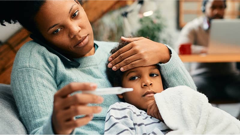 A mother holds a thermometer and checks her child’s forehead while the child rests under a blanket on the couch.