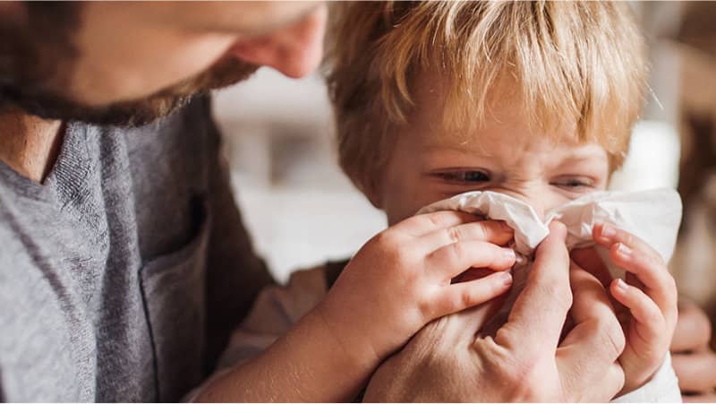 A father helps a young child wipe their nose with a tissue while the child looks upset.