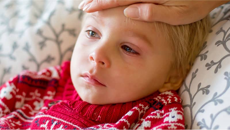 A sick child in a red sweater lies on a bed while an adult hand rests on their forehead to check for fever.