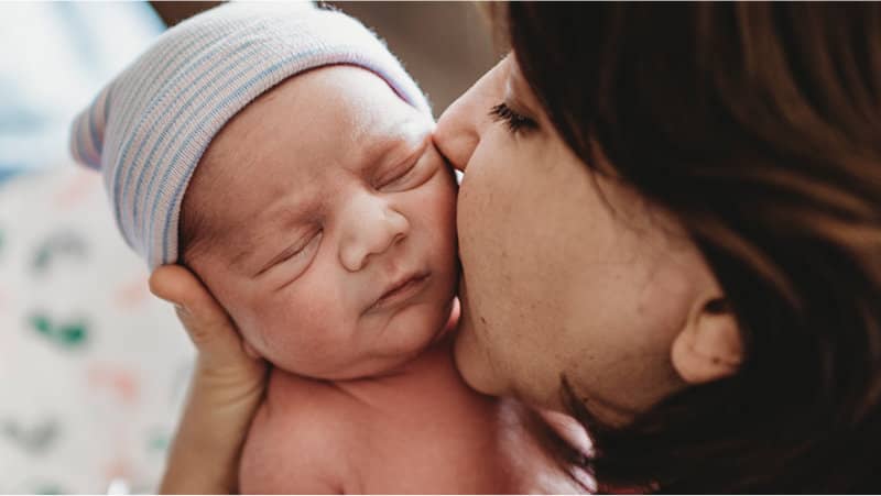 A mother kisses a sleeping newborn wearing a striped hat while gently supporting the baby’s head.