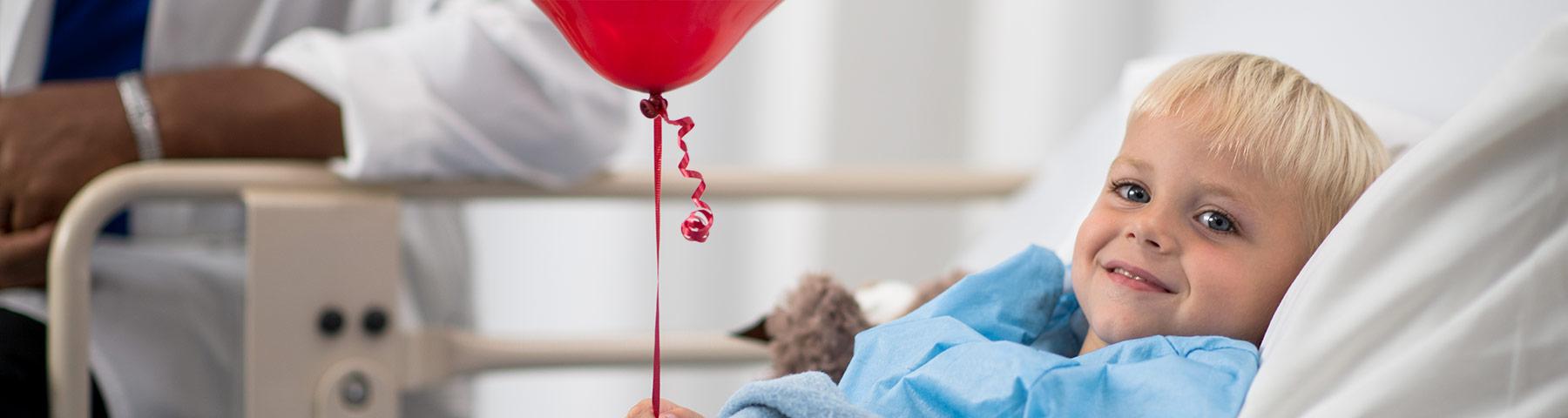Child laying in bed smiling holding a red balloon with his doctor at bedside.