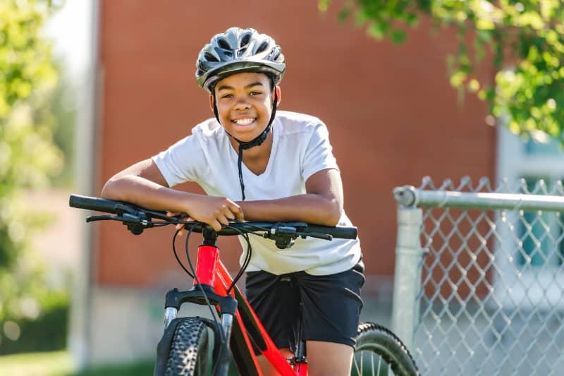 Smiling child wearing a bike helmet sitting on a red bicycle outdoors near a chain-link fence and school building.