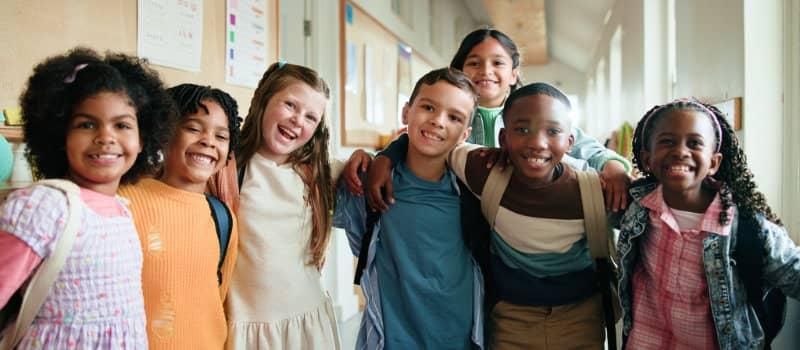 Group of diverse elementary school children smiling with their arms around each other in a bright school hallway.