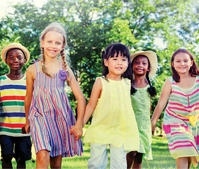 Five young children of diverse backgrounds holding hands and smiling while walking together outdoors.