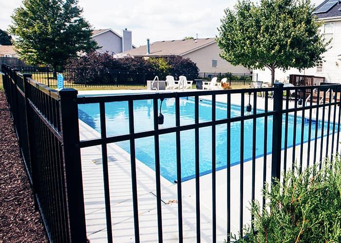 A fenced-in outdoor swimming pool in a residential neighborhood with chairs and trees around it, emphasizing pool safety with a secure gate.