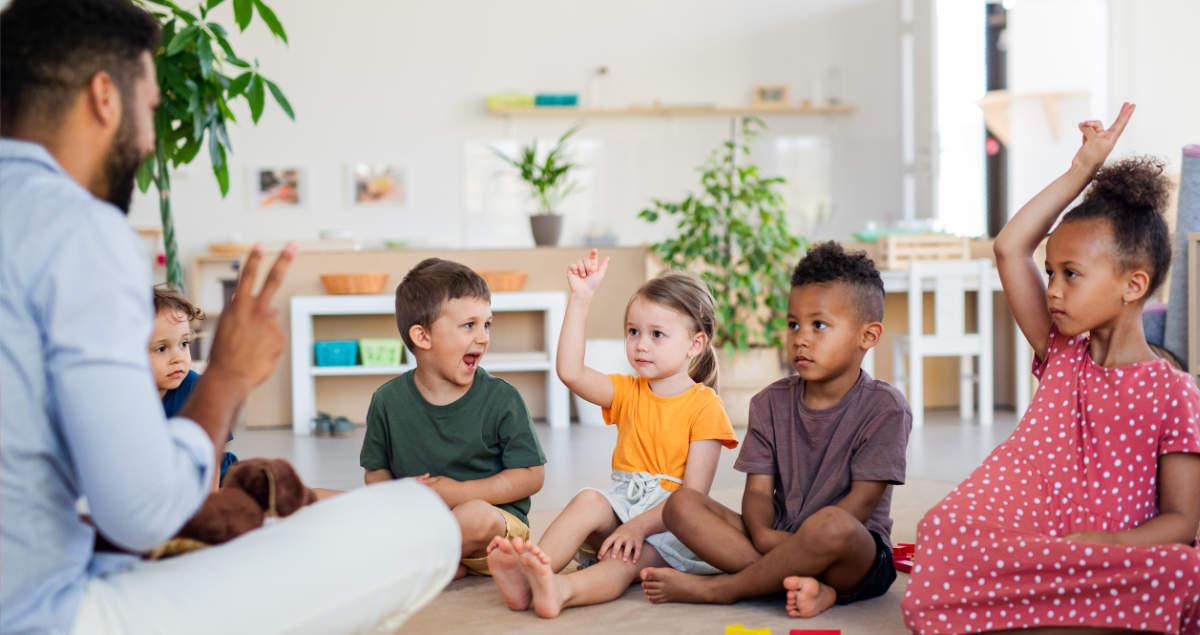 A teacher sits on the floor facing four young children in a bright classroom, with several children raising their hands to participate. Indoor plants and colorful shelves are visible in the background.