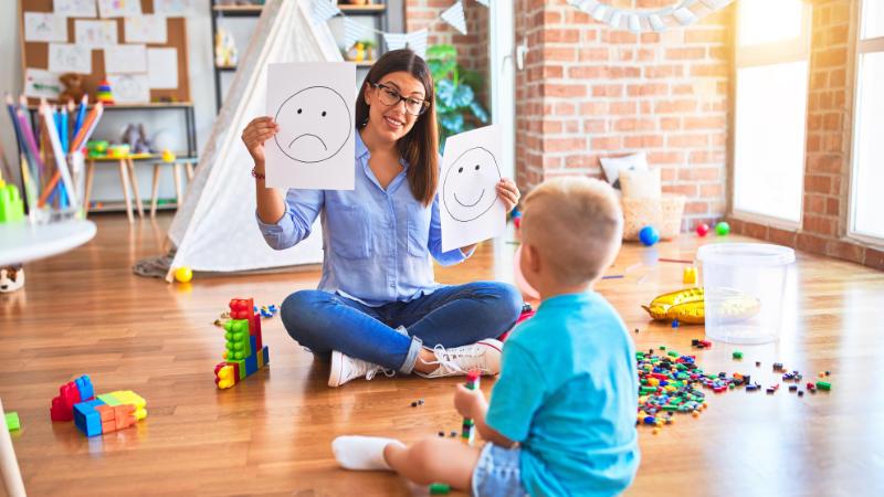 A smiling female teacher sits cross-legged on a wooden floor with a young boy, holding up two cards with sad and happy faces to teach about emotions in a colorful classroom.